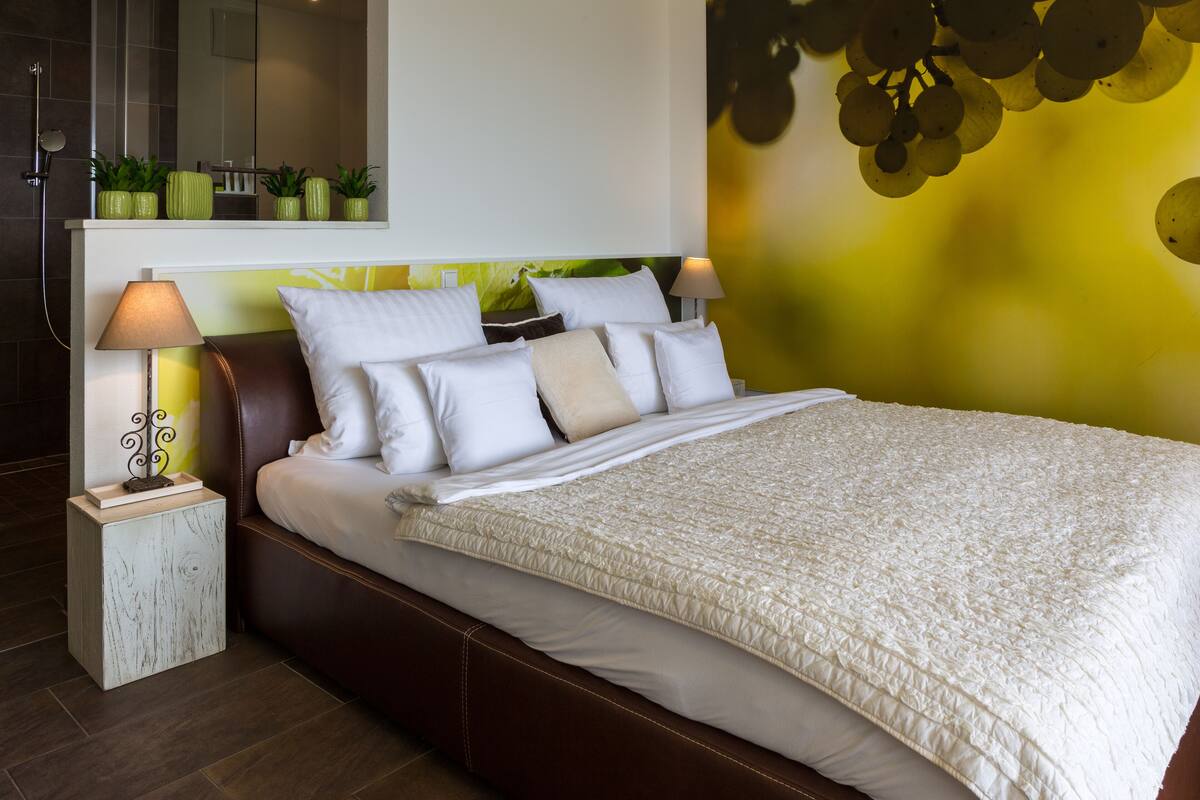 A neatly made bed with white linens and a beige quilt, a bedside table with a lamp, and a wall with a green and yellow abstract design.