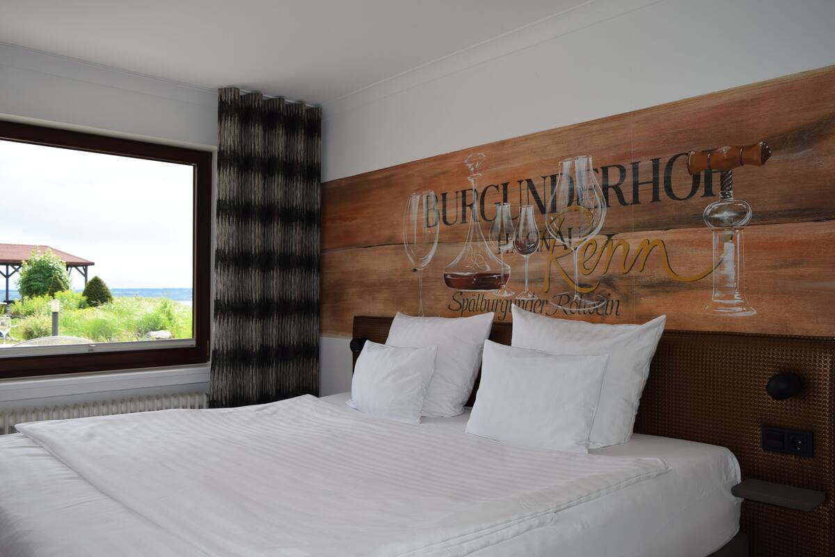 A hotel room with a large bed, white bedding, a view of the outdoors through a window, and a wooden wall with a mural of wine glasses and text.