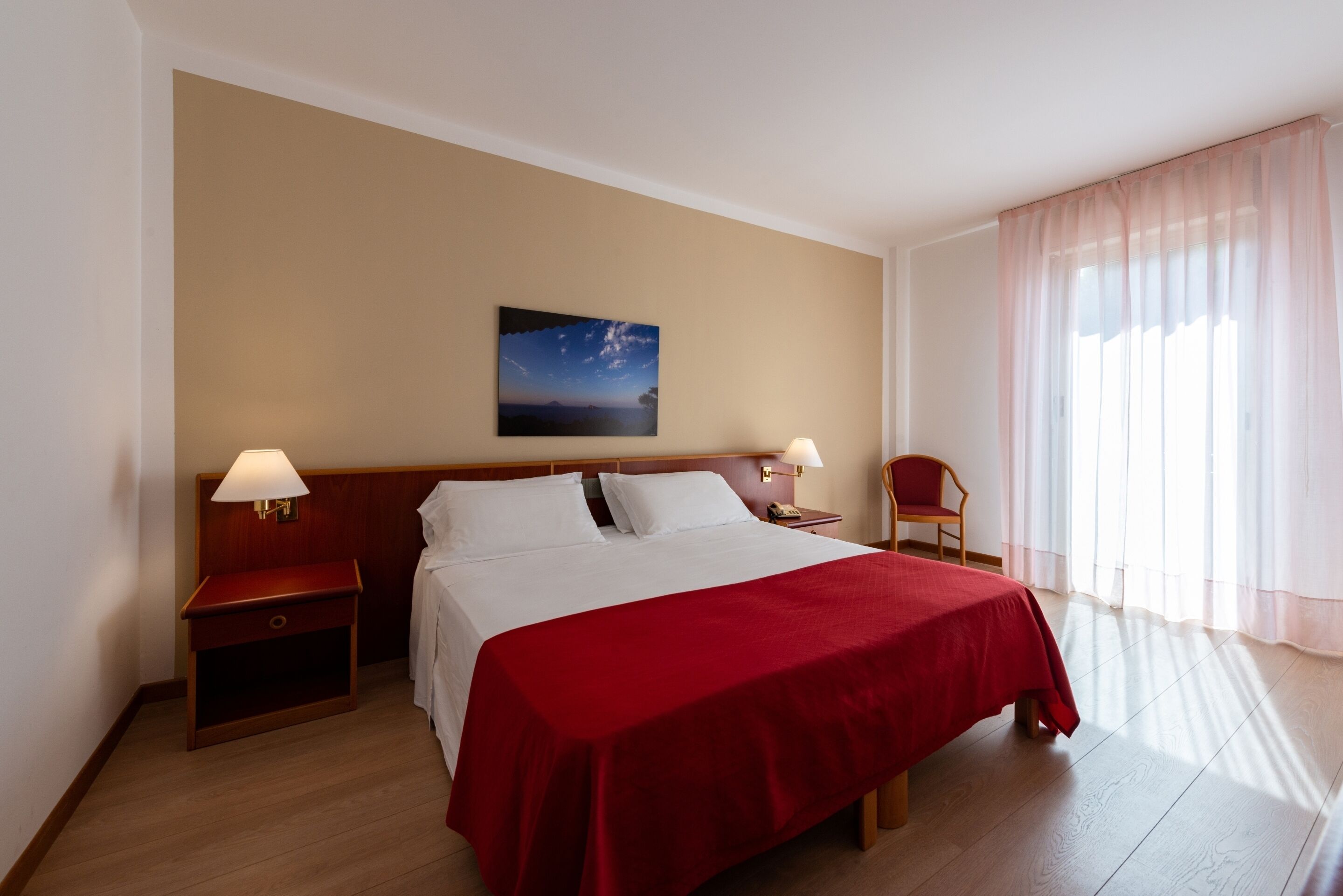 Una camera d'albergo con un letto, un comodino, una sedia, una lampada e una finestra con tende.
