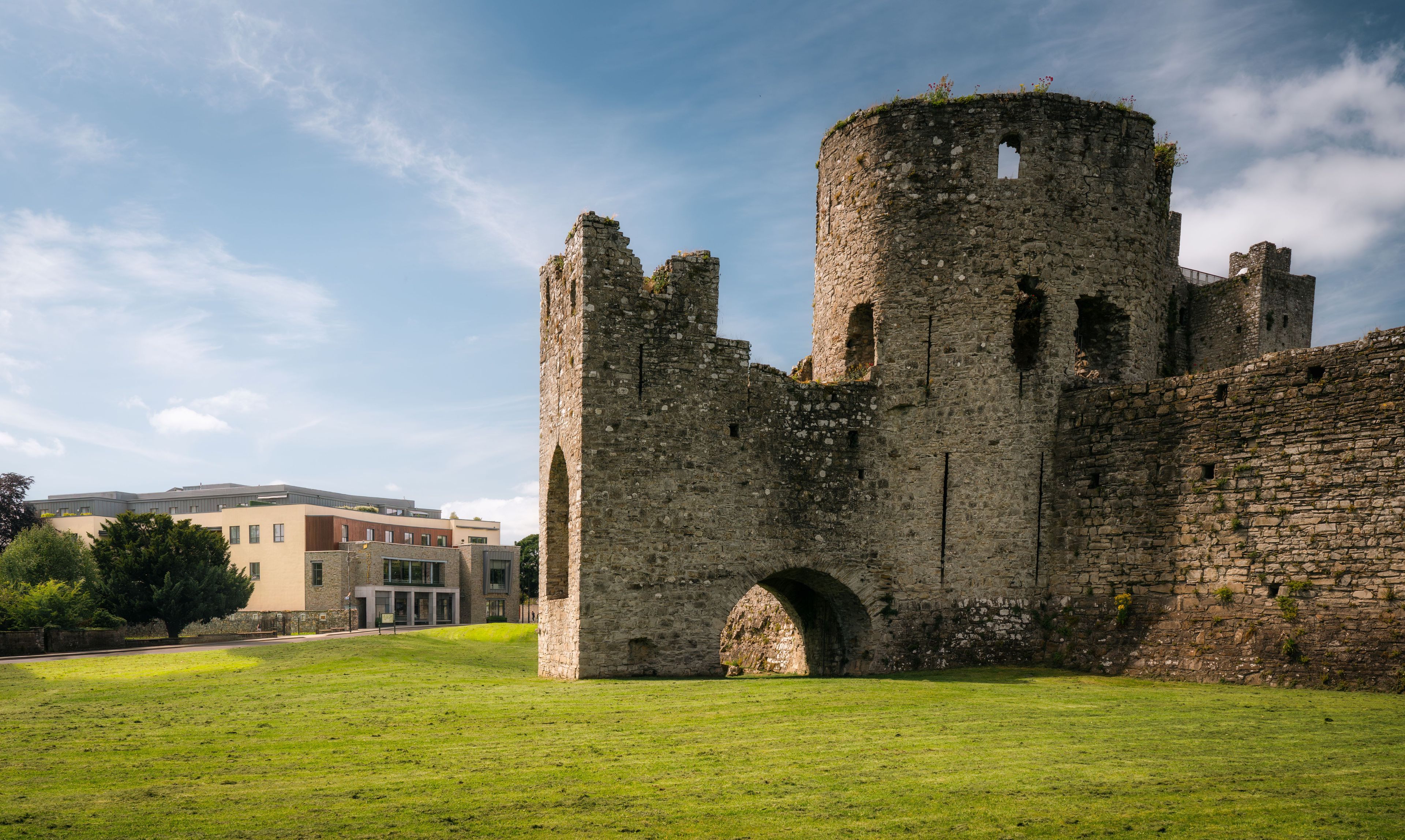 A historic stone castle with a large archway and a round tower, surrounded by a grassy field and modern buildings in the background.