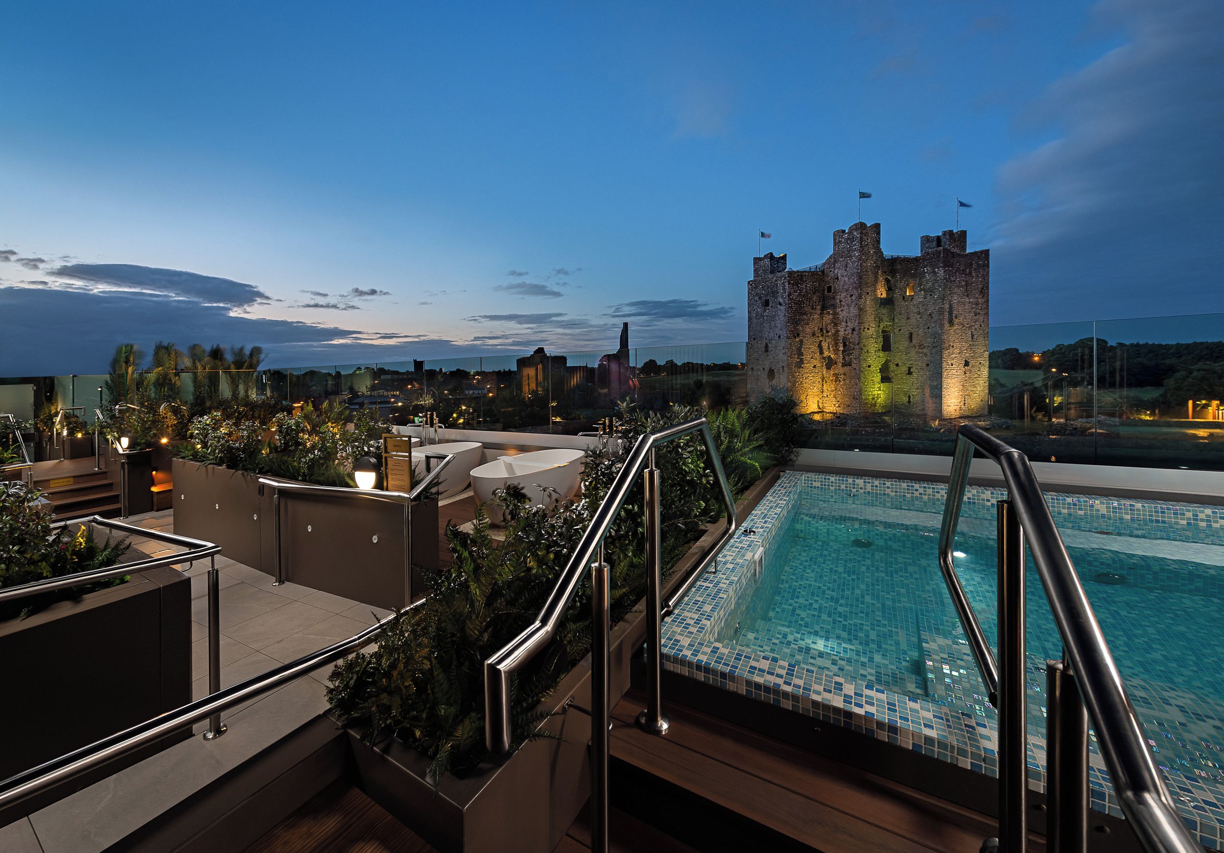 A rooftop pool with a castle in the background, surrounded by greenery and modern buildings.