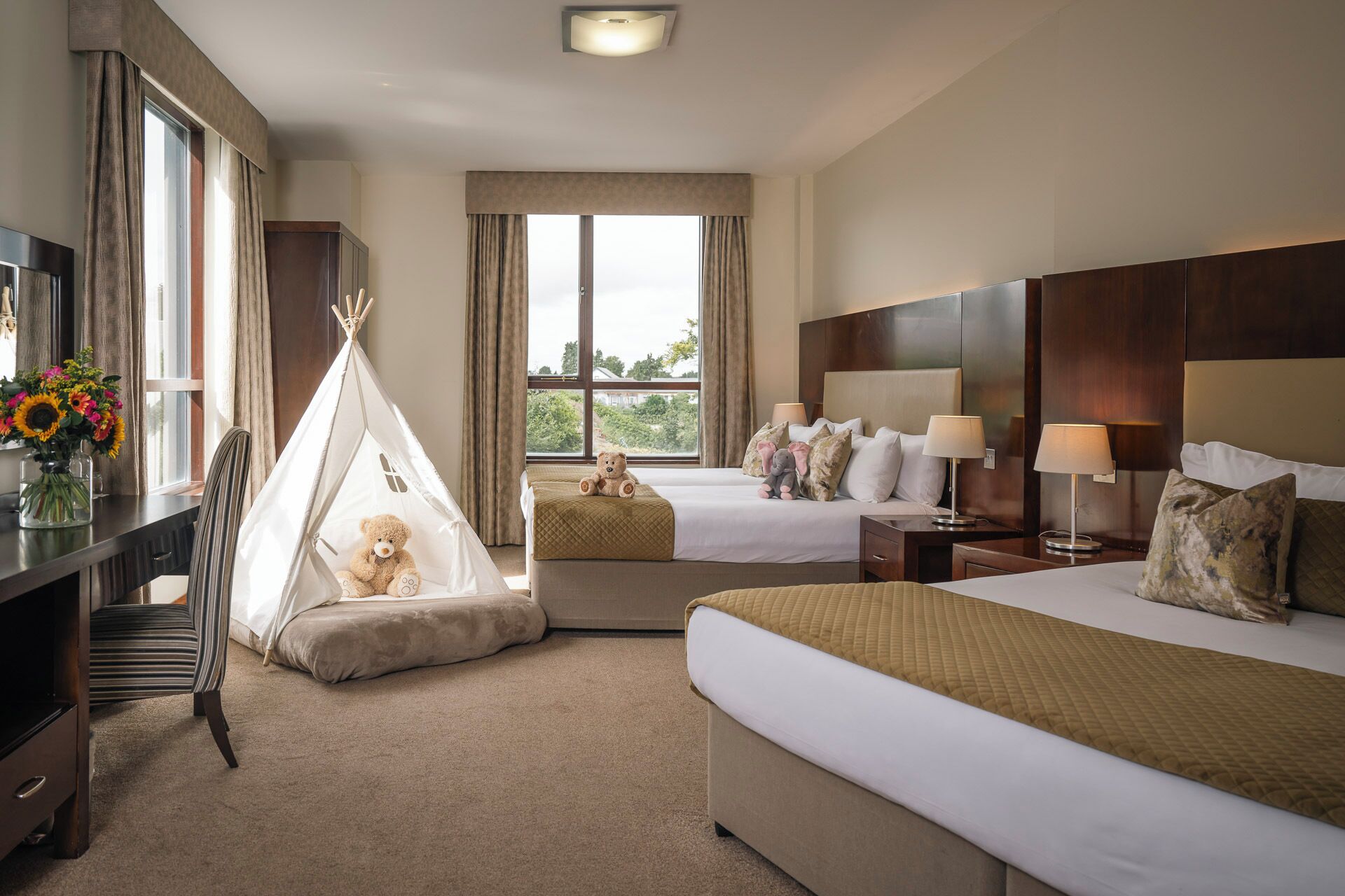 A hotel room with two beds, a desk, a chair, a TV, a window with curtains, and a teddy bear in a teepee.