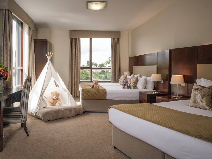 A hotel room with two beds, a desk, a chair, a TV, a window with curtains, and a teddy bear in a teepee.