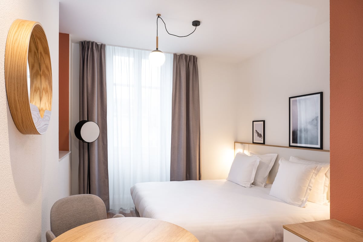 A hotel room with a bed, a desk, a chair, a round wooden table, a round mirror, a hanging light fixture, and a window with curtains.