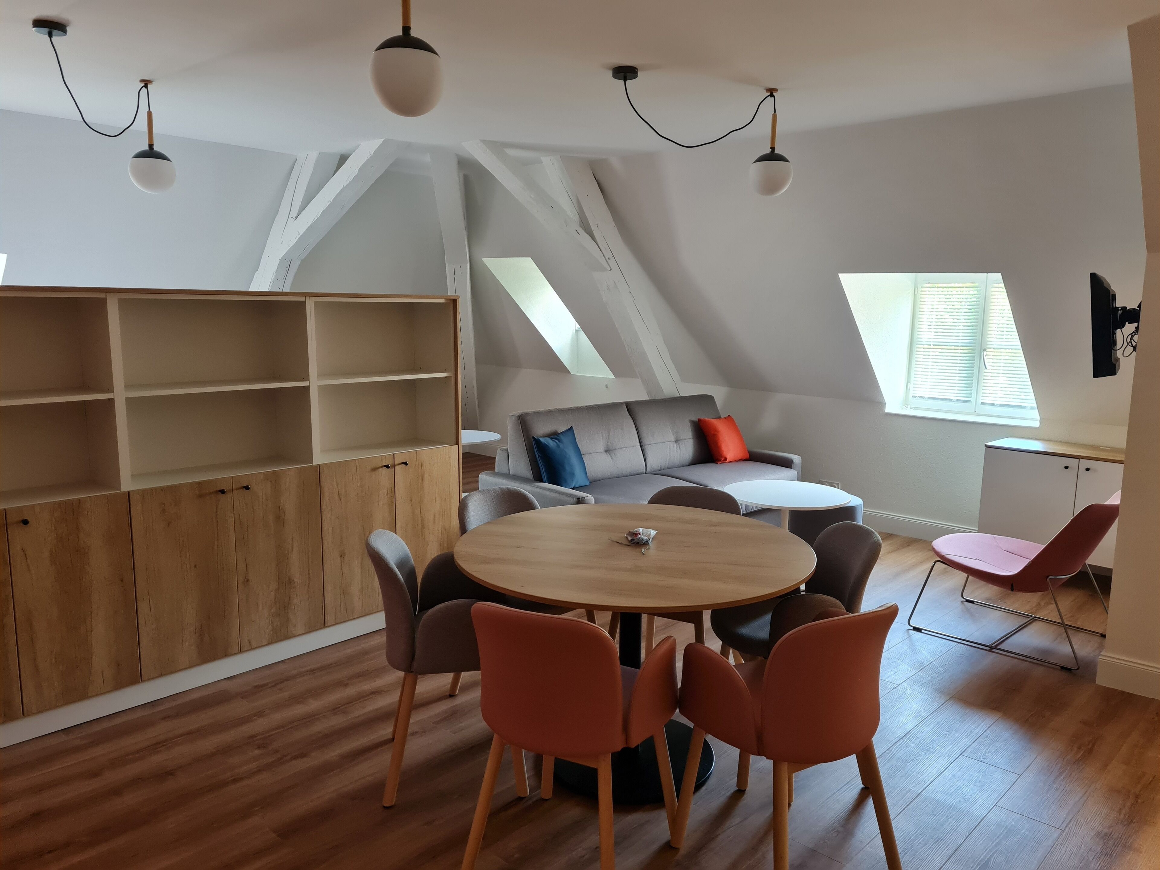 Een moderne loftkamer met een houten eettafel, oranje stoelen, een grijze bank met rode en blauwe kussens en een witte kast.