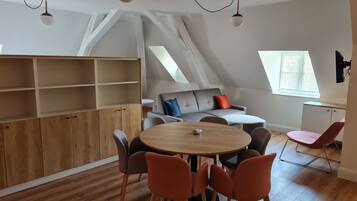 Een moderne loftkamer met een houten eettafel, oranje stoelen, een grijze bank met rode en blauwe kussens en een witte kast.