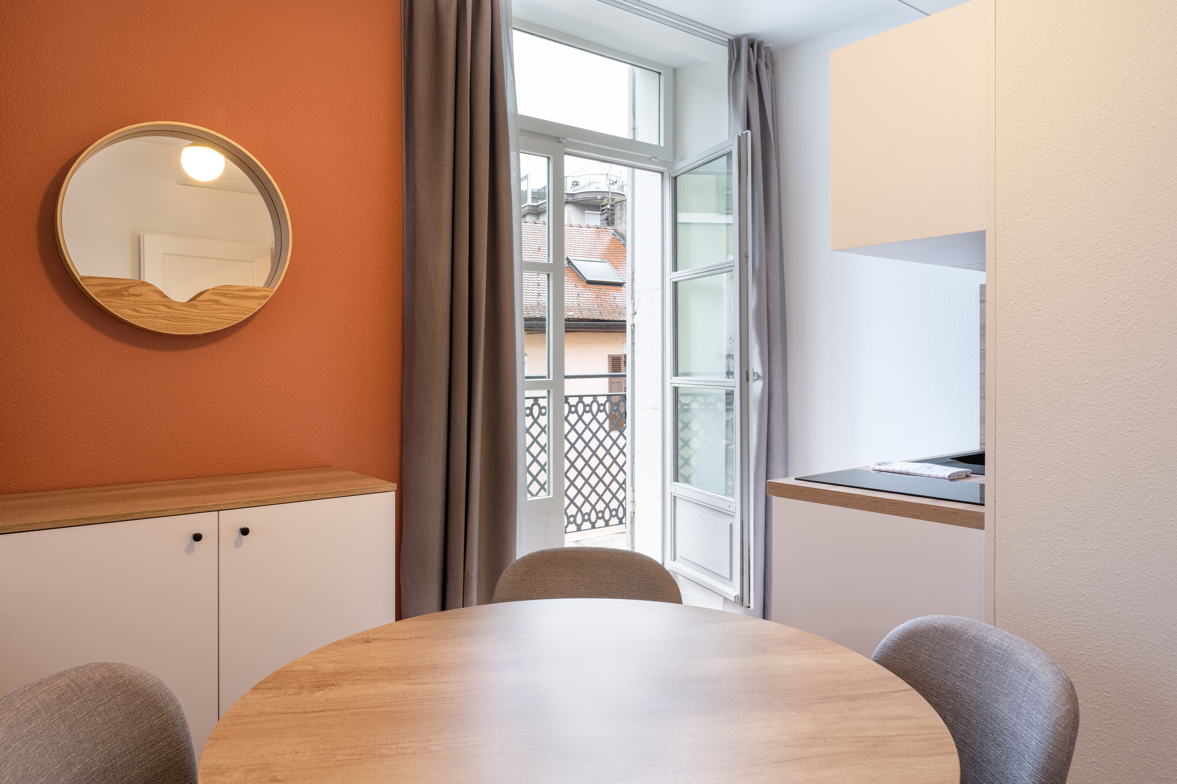 Ein Zimmer mit einem runden Holztisch, zwei Stühlen, einem Rundspiegel, einem Fenster mit Vorhängen und Blick auf Gebäude.