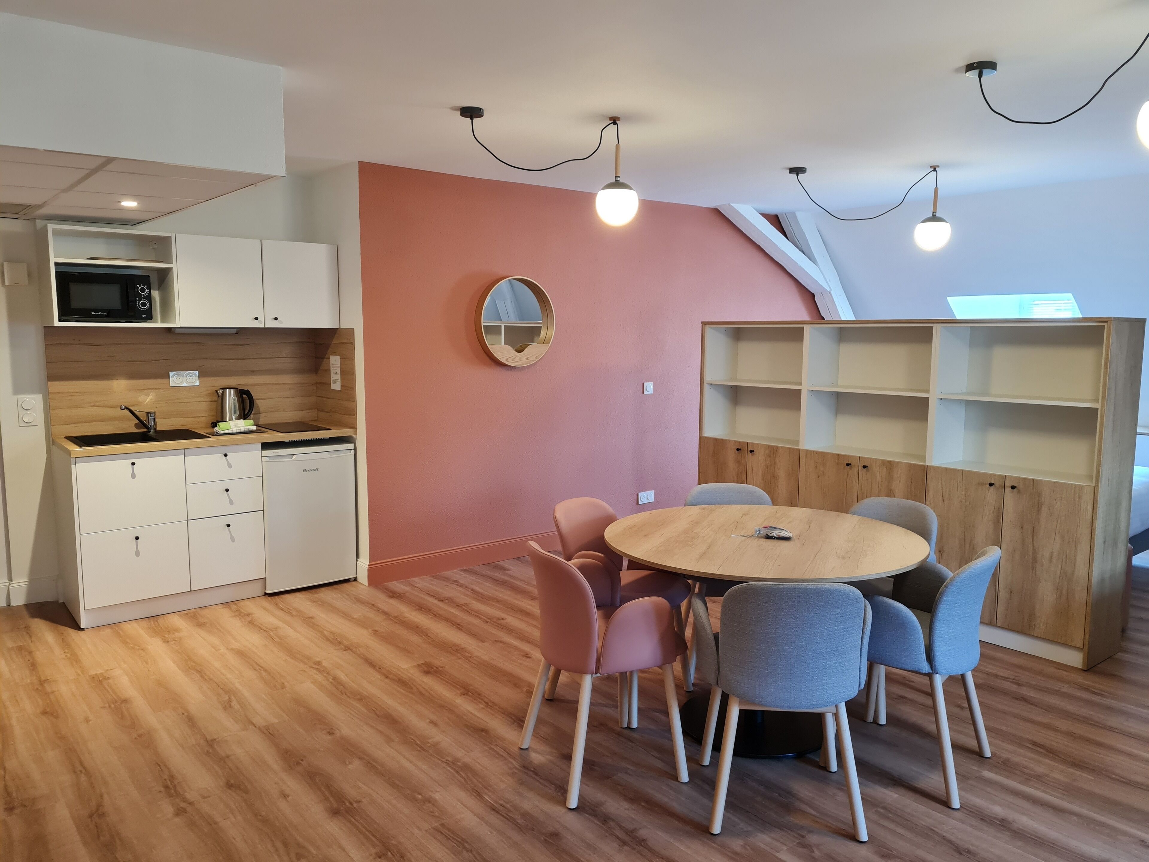 Een moderne keuken met witte kasten, een magnetron en een spoelbak. Aan de muur hangt een ronde spiegel en er staat een houten eettafel met stoelen.