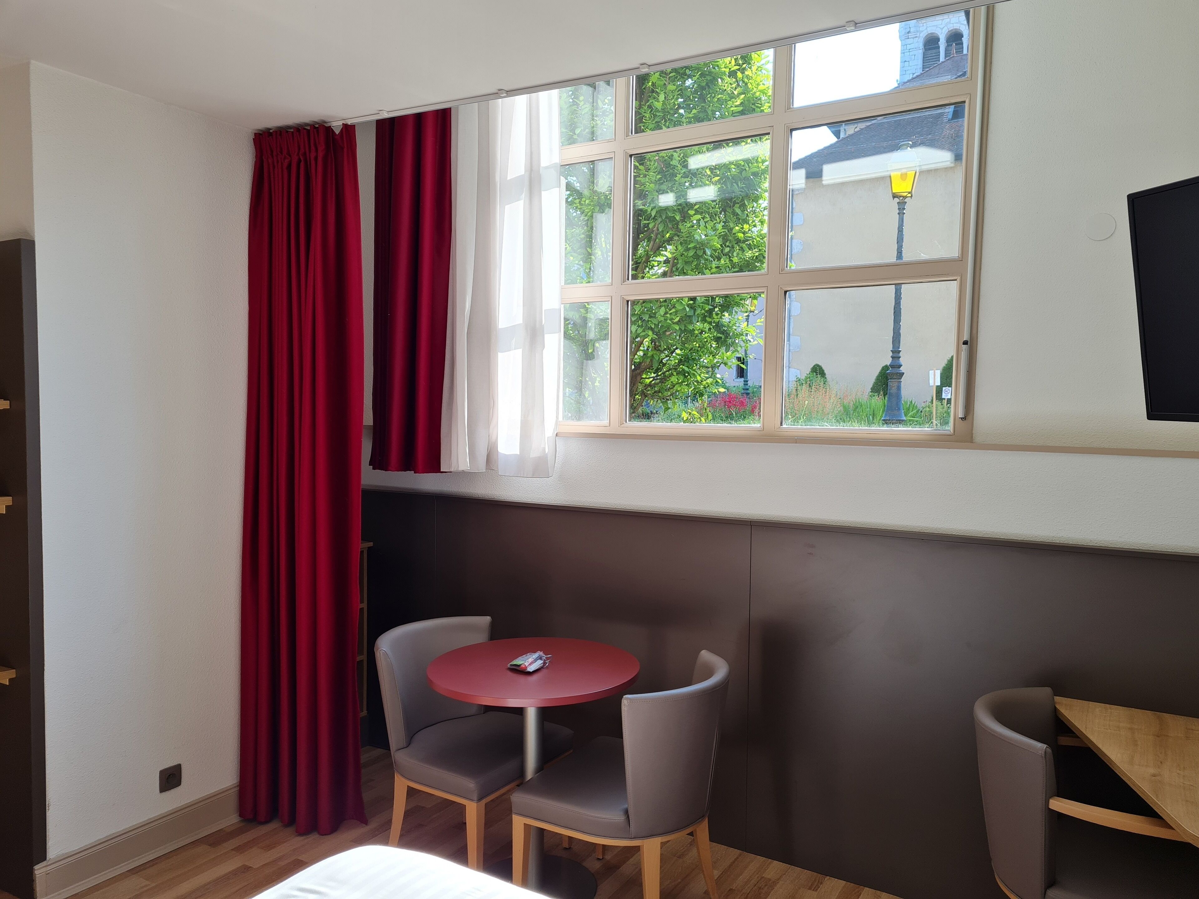 Ein Zimmer mit Fenster, einem roten Tisch, zwei Stühlen und einem Bett.