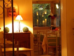 A warmly lit dining area with a table set for two, a lamp, and decorative plants. - Villa Glanum (St.-Rémy-de-Provence)