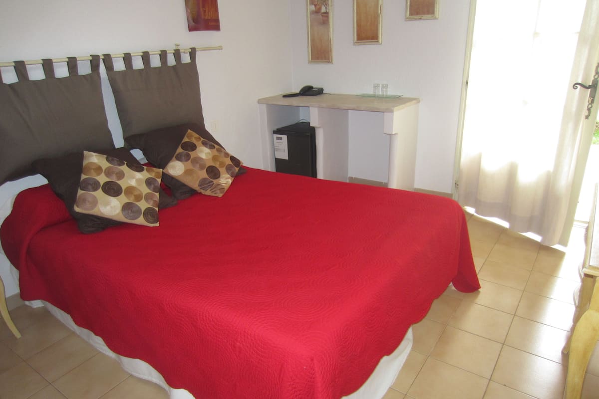A bed with a red bedspread and two patterned pillows, a bedside table with a phone and glasses, and a window with curtains.