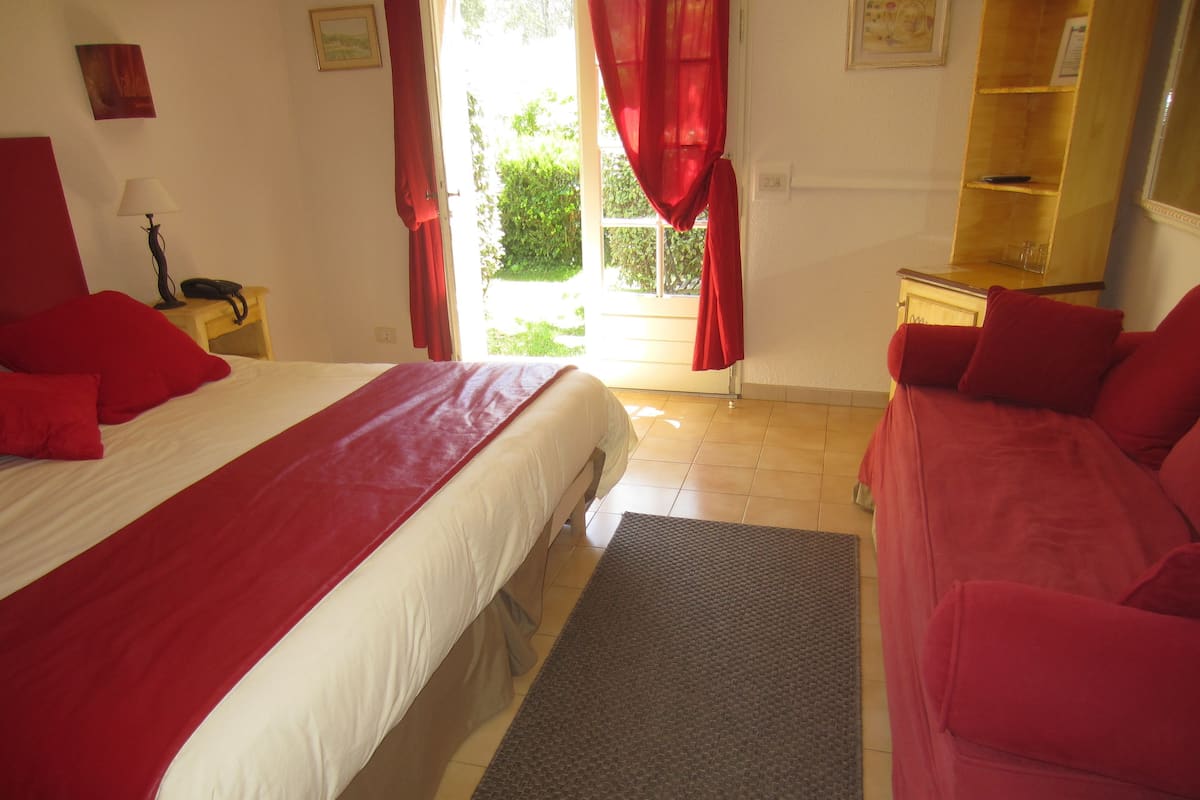A hotel room with a large bed, a sofa, a small table, and a window with red curtains.