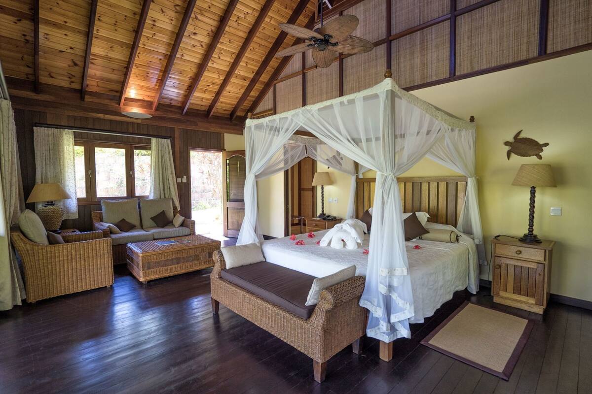 A room with a canopy bed, wicker furniture, a wooden ceiling, and a view through the window.