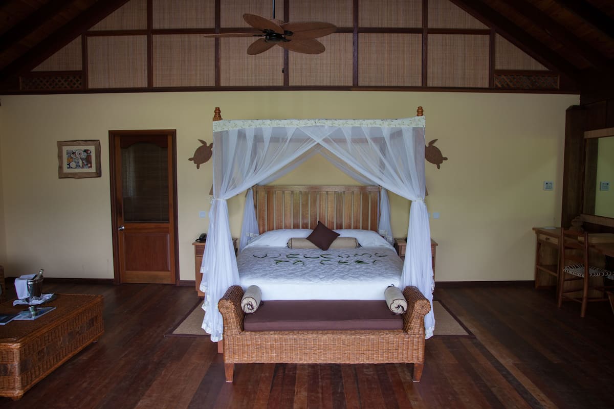 A spacious bedroom with a four-poster bed, a wooden ceiling fan, and a wooden floor.