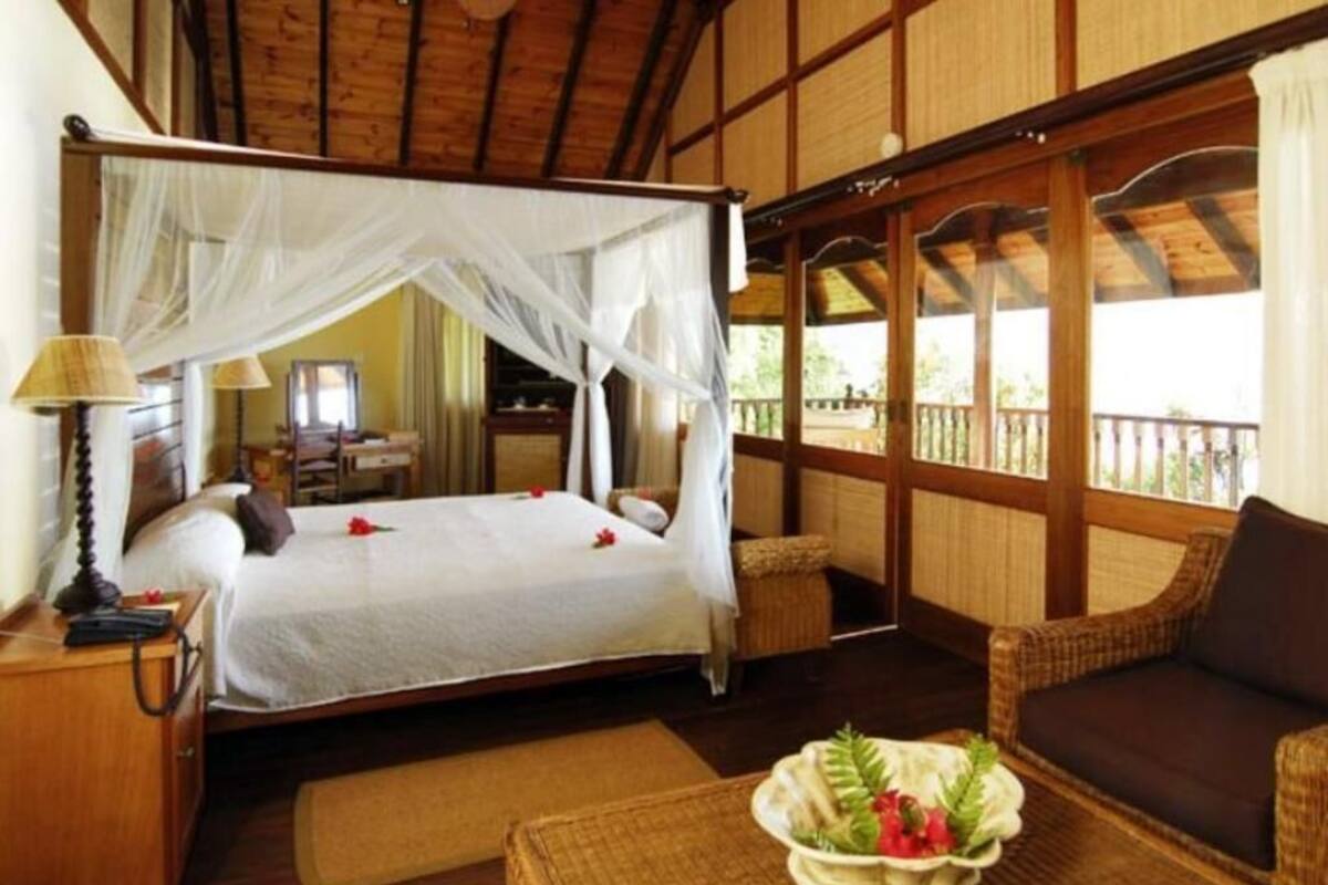 A room with a bed under a canopy, a wicker sofa, a small table with a decorative bowl, and a wooden ceiling.