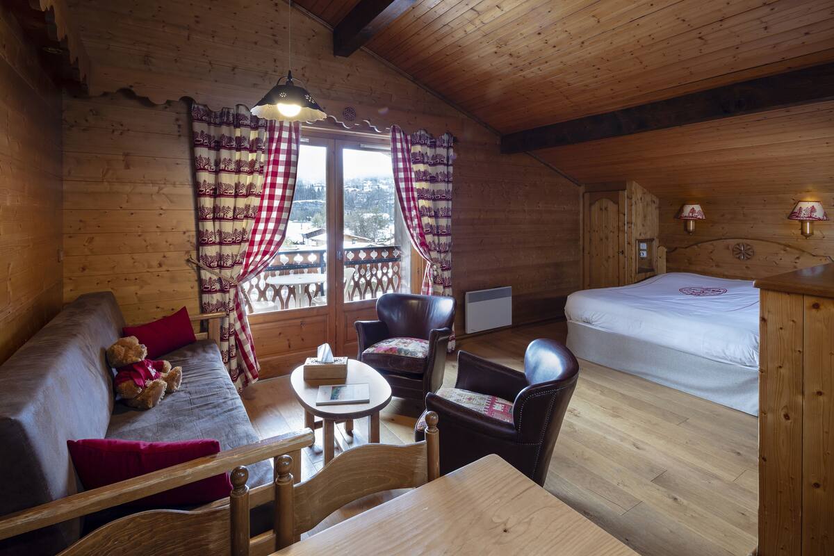 A cozy wooden cabin room with a bed, a sofa, a chair, a table, and a window with curtains.
