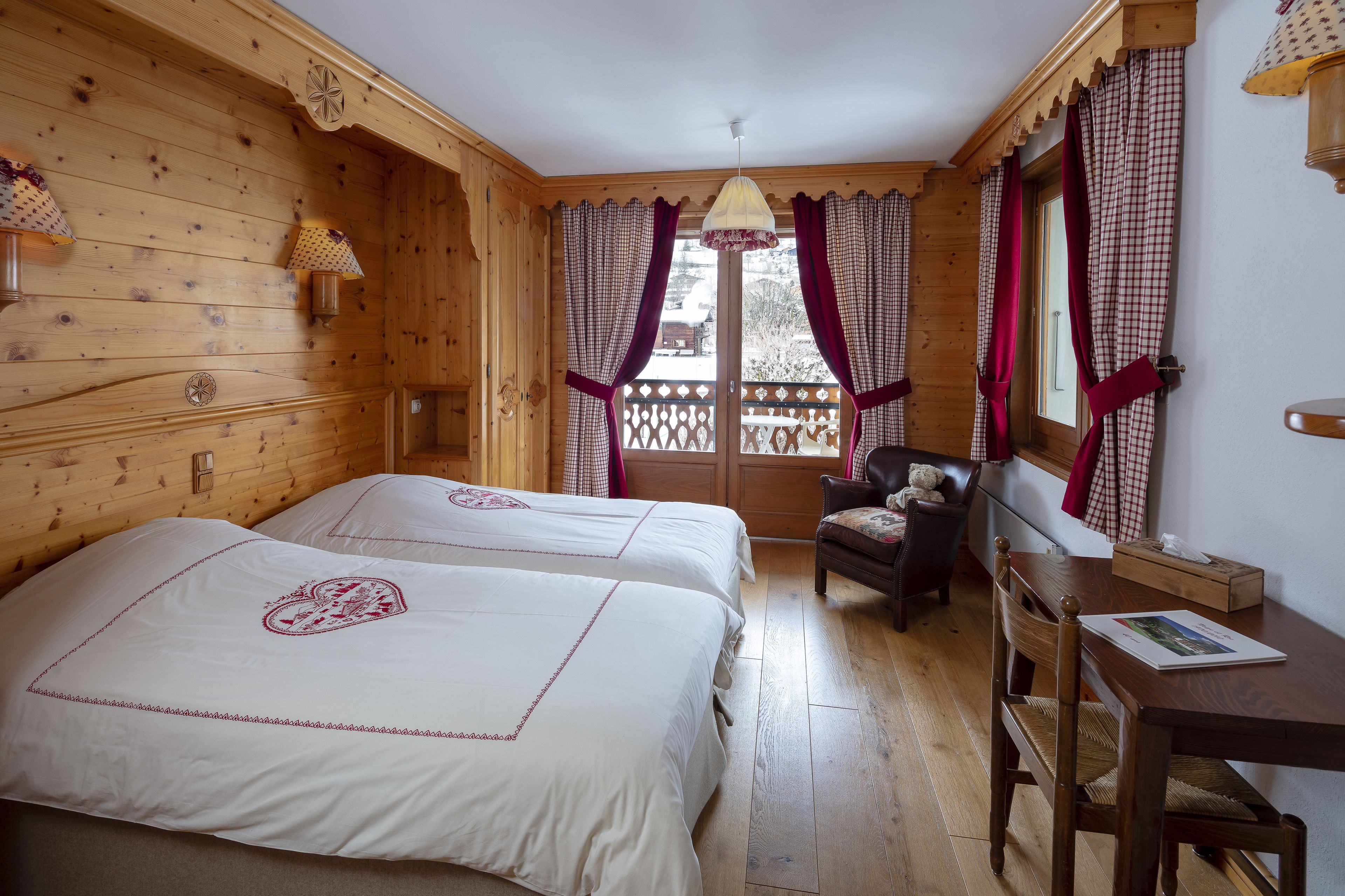Ein Zimmer mit Holzboden, zwei Betten, einem Sessel, einem Tisch und einem Fenster mit Vorhängen.