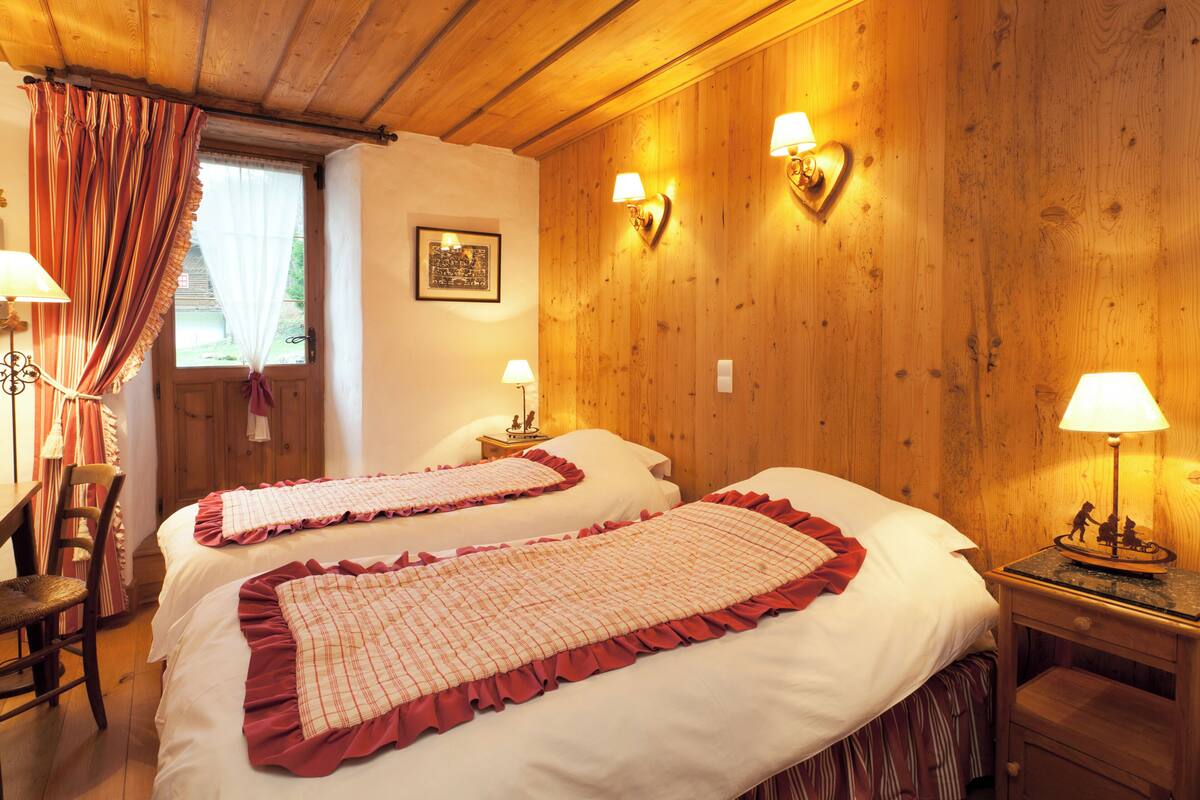 A room with two beds, wooden walls, a window with curtains, and wall-mounted lamps.