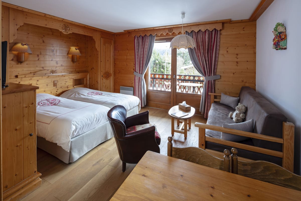 A wooden cabin room with two beds, a sofa, a table, and a window with curtains.