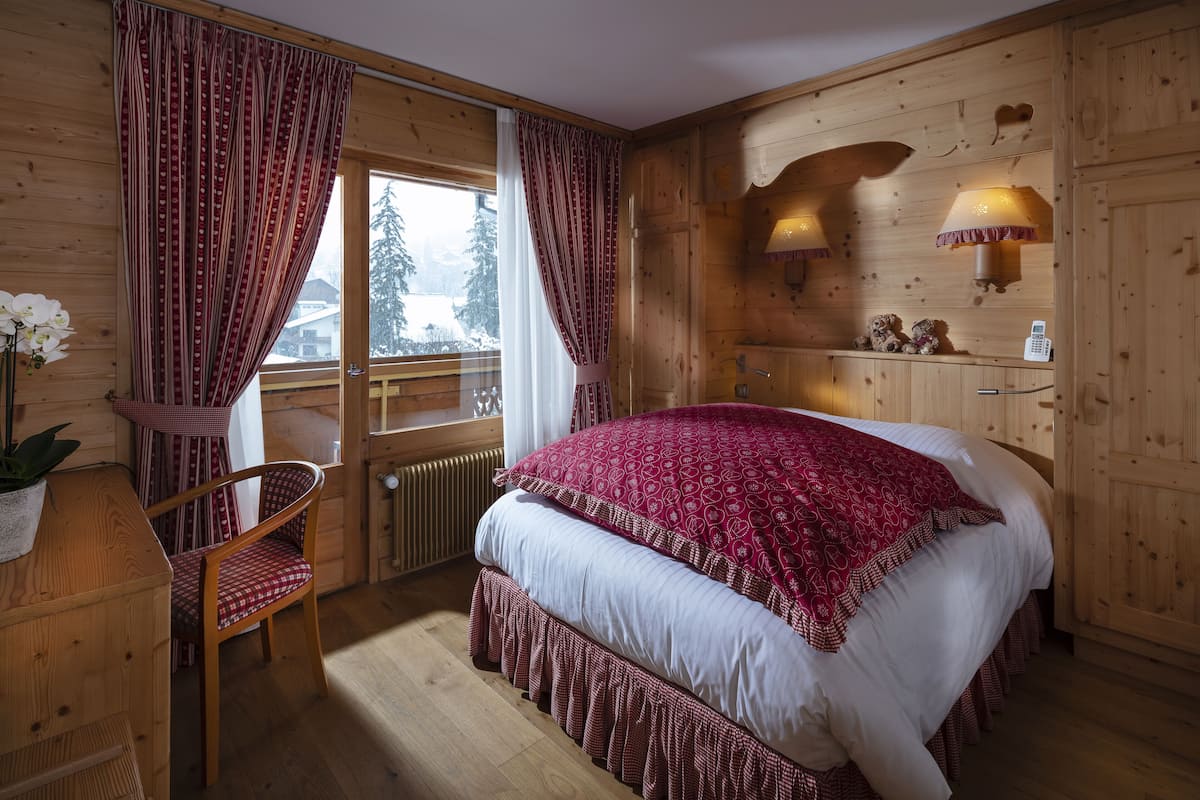 A wooden cabin bedroom with a large bed, a chair, a window with curtains, a lamp, and a shelf with decorative items.