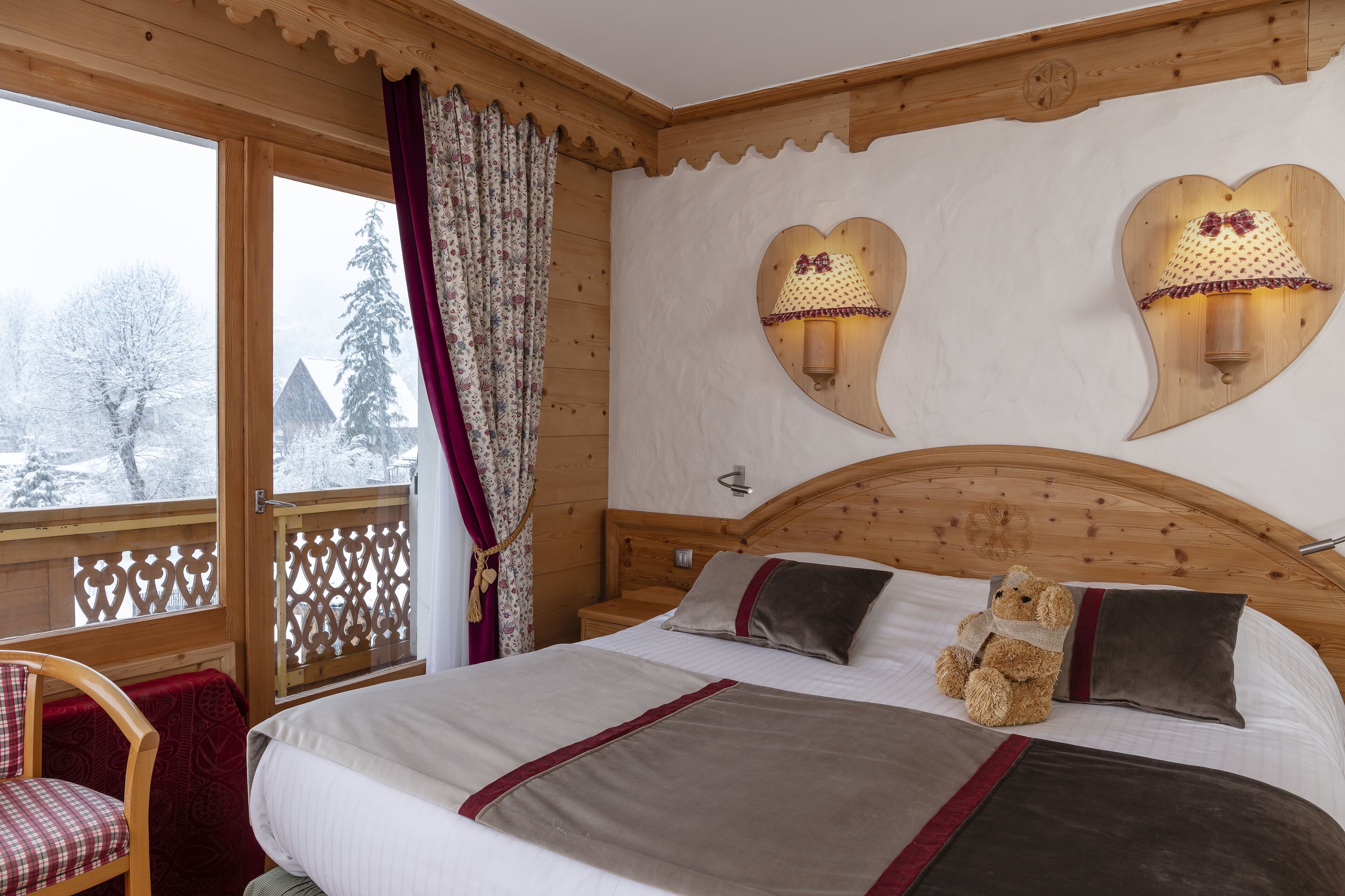 A cozy wooden cabin bedroom with a large bed, two heart-shaped wall sconces, a chair, and a view of a snowy landscape.