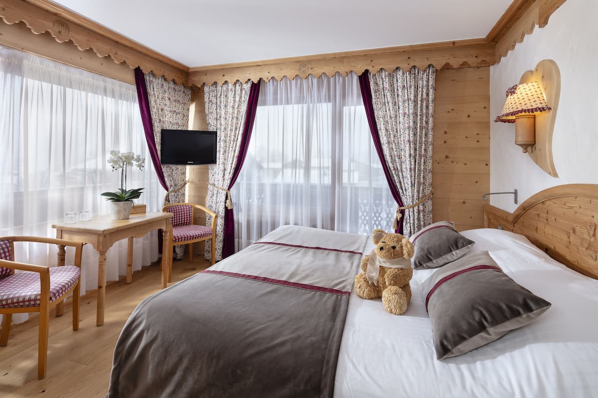 A hotel room with a bed, a desk with a chair, a television, and a teddy bear on the bed.