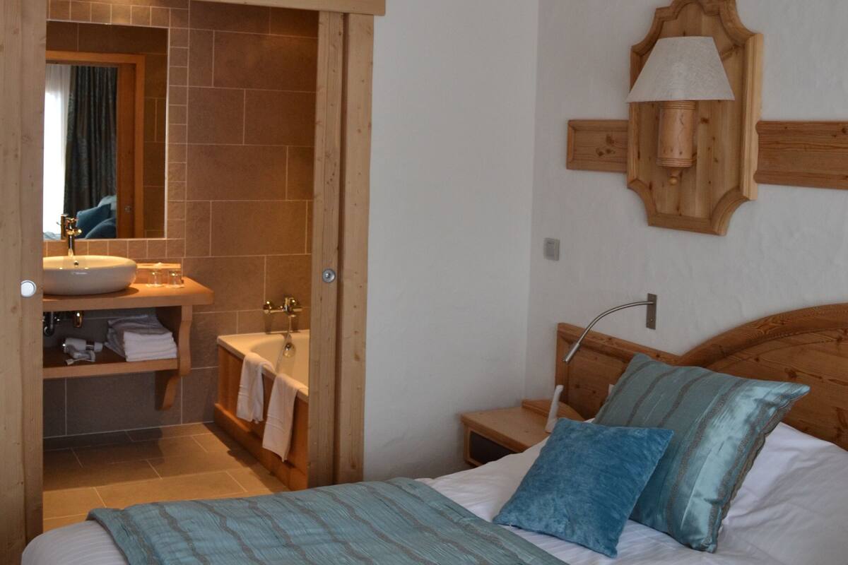 A bedroom with a bed, a wooden headboard, a nightstand, a bedside table, a mirror, and a bathroom visible through an open door.