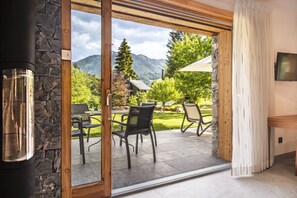 A room with a glass door leading to an outdoor patio with chairs and a table, a stone wall, and a television on the wall. - Neige et Roc, The Originals Relais (Samoëns)