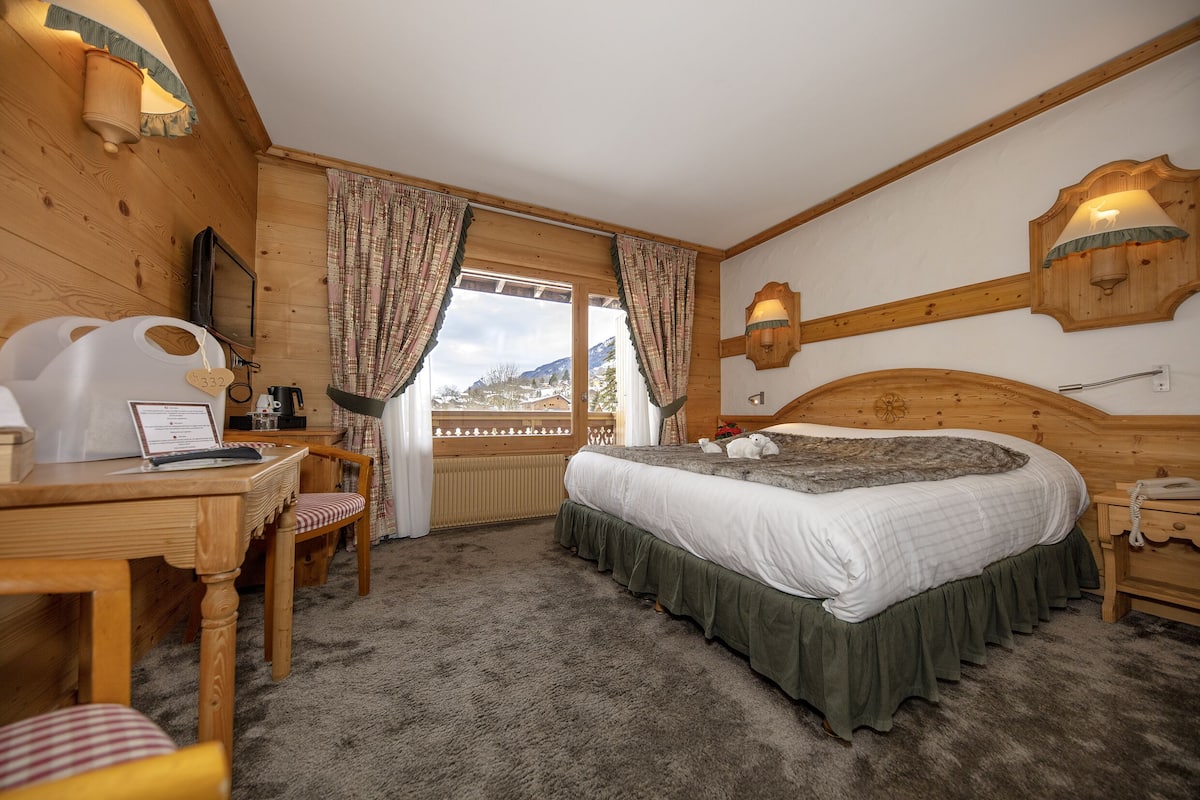 A cozy room with a wooden bed, a desk with a laptop, and a view of mountains through the window.