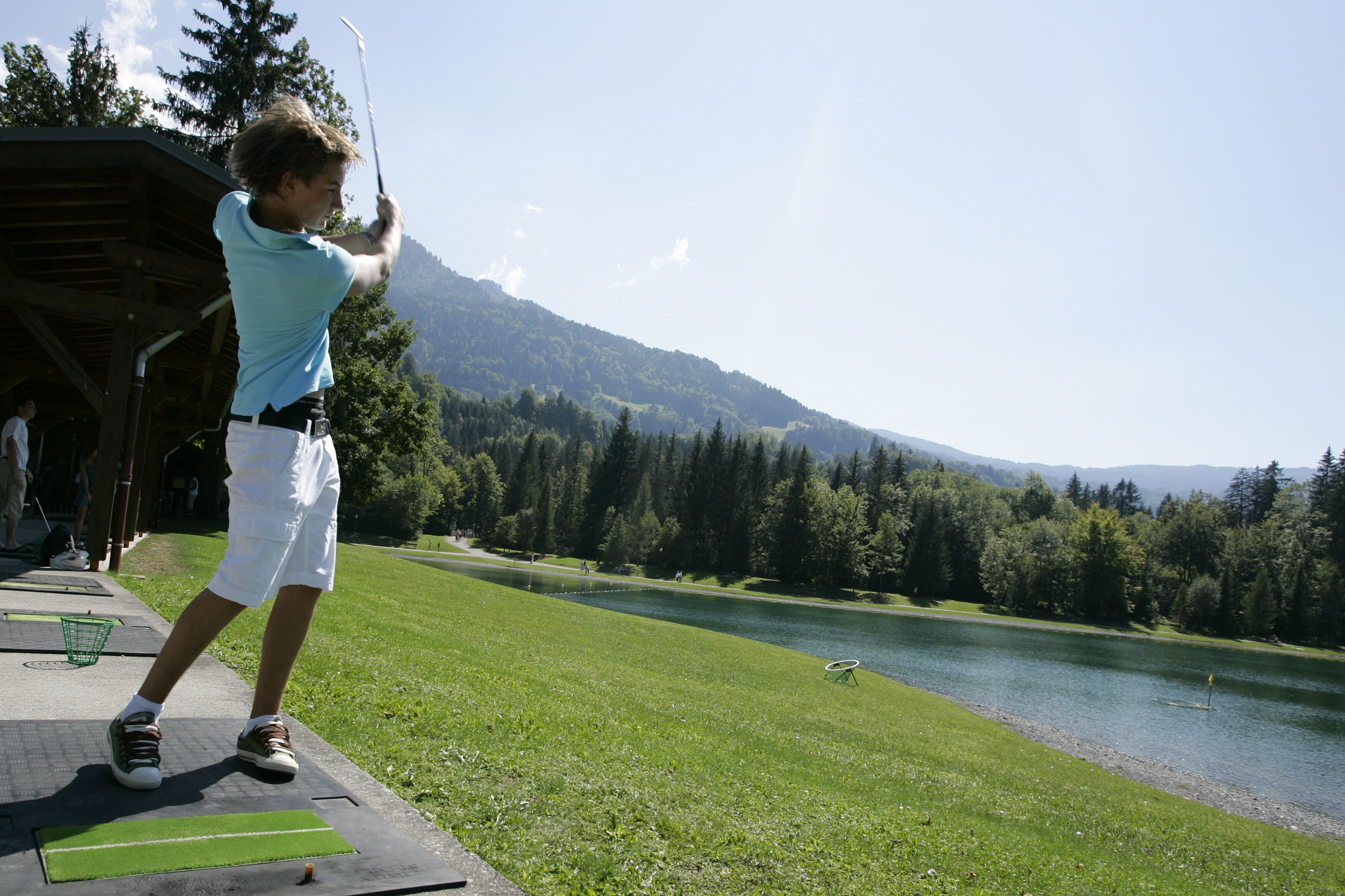 A person playing golf on a course with a lake and forested hills in the background.