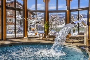 A pool area with a water feature, lounge chairs, and a view of a snowy landscape through large windows. - Neige et Roc, The Originals Relais (Samoëns)