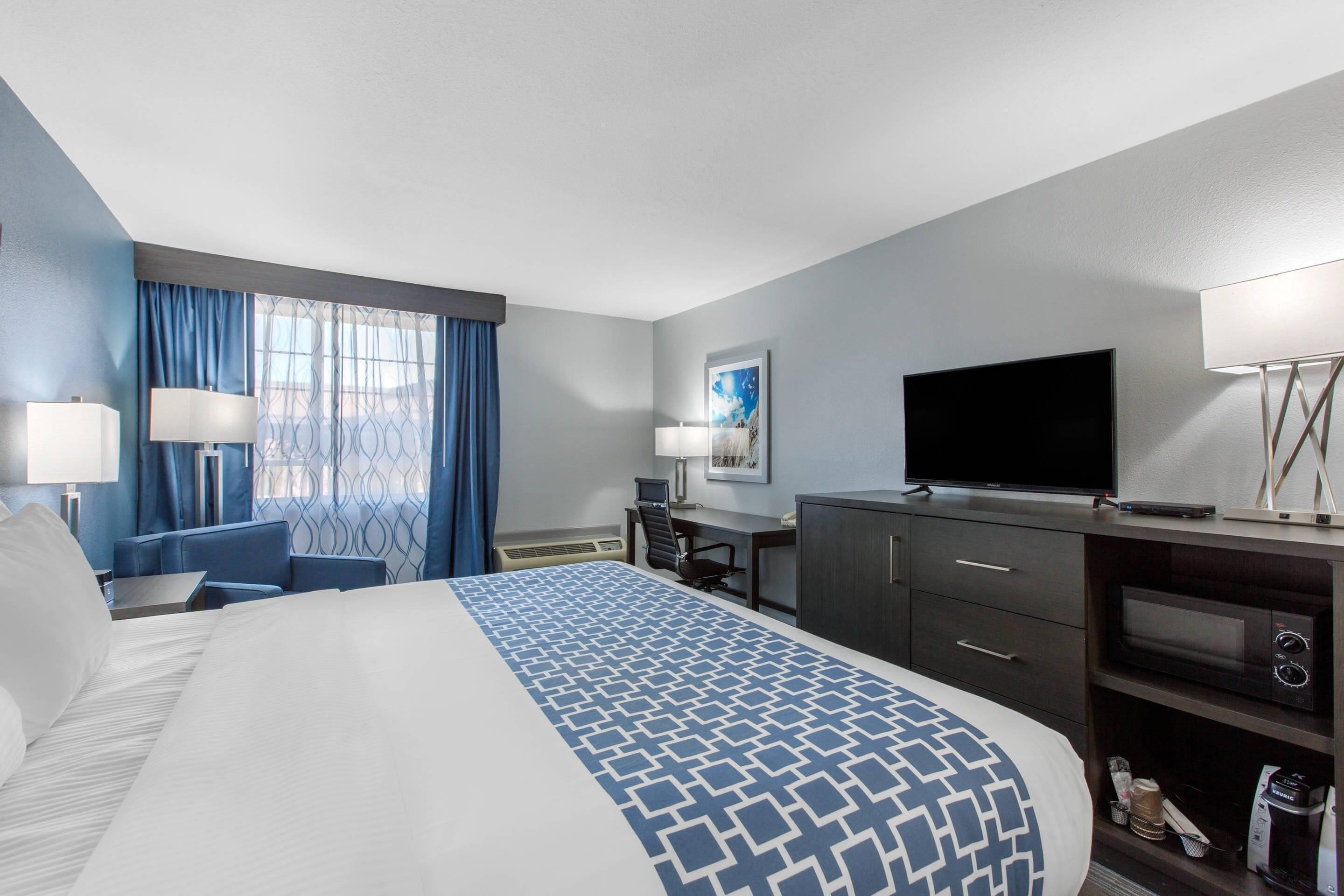 A hotel room with a large bed, a desk, a TV, and a minibar.
