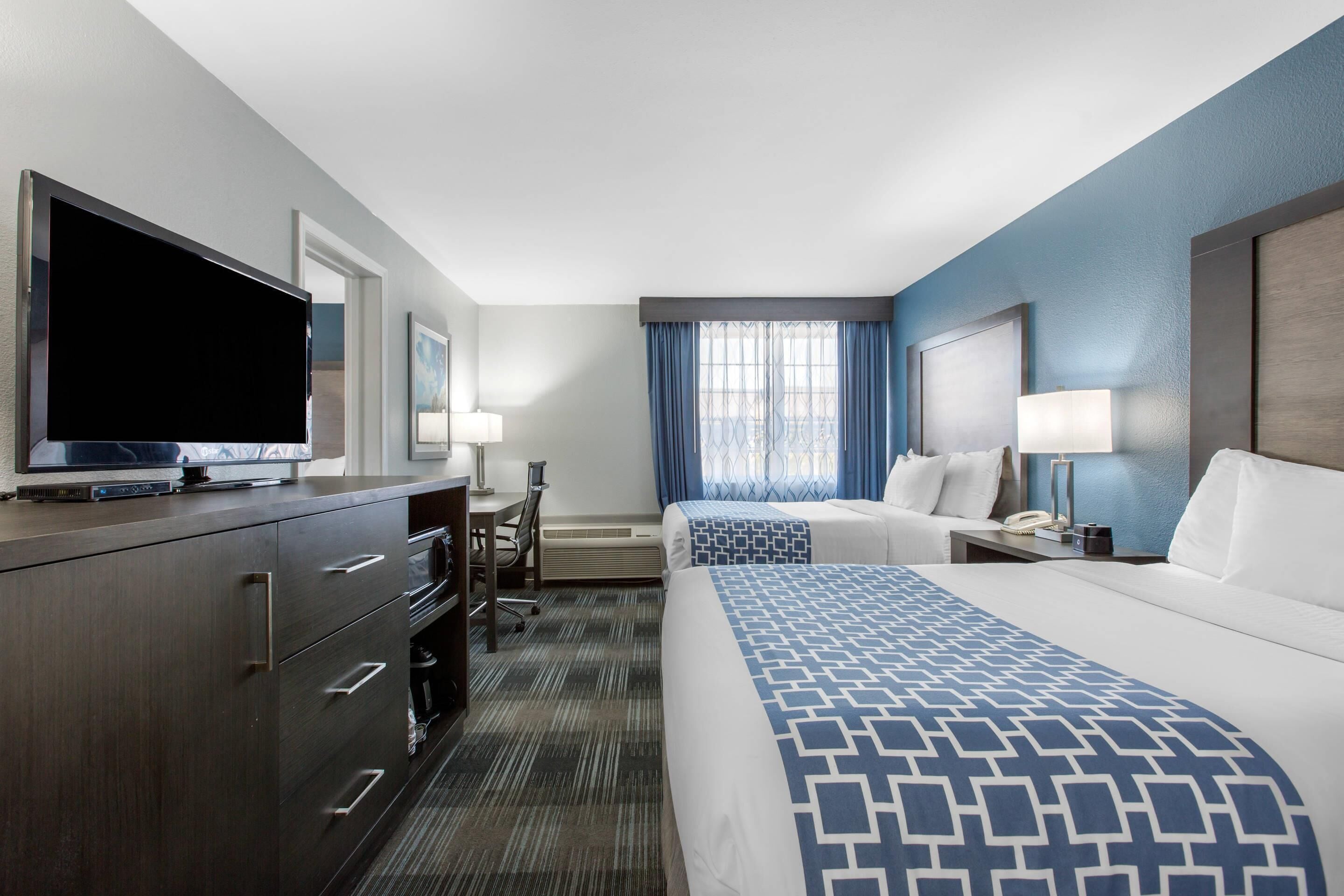 A hotel room with a large flat-screen TV, a desk with a lamp, two beds with blue and white bedding, and a window with blue curtains.