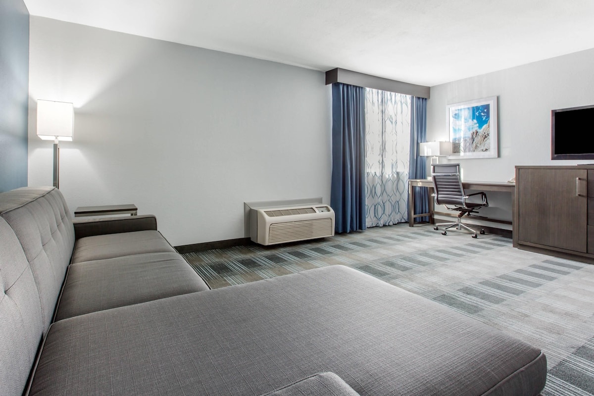 Une chambre d’hôtel moderne avec un canapé gris, un bureau avec une chaise, un appareil de climatisation et un téléviseur fixé au mur.