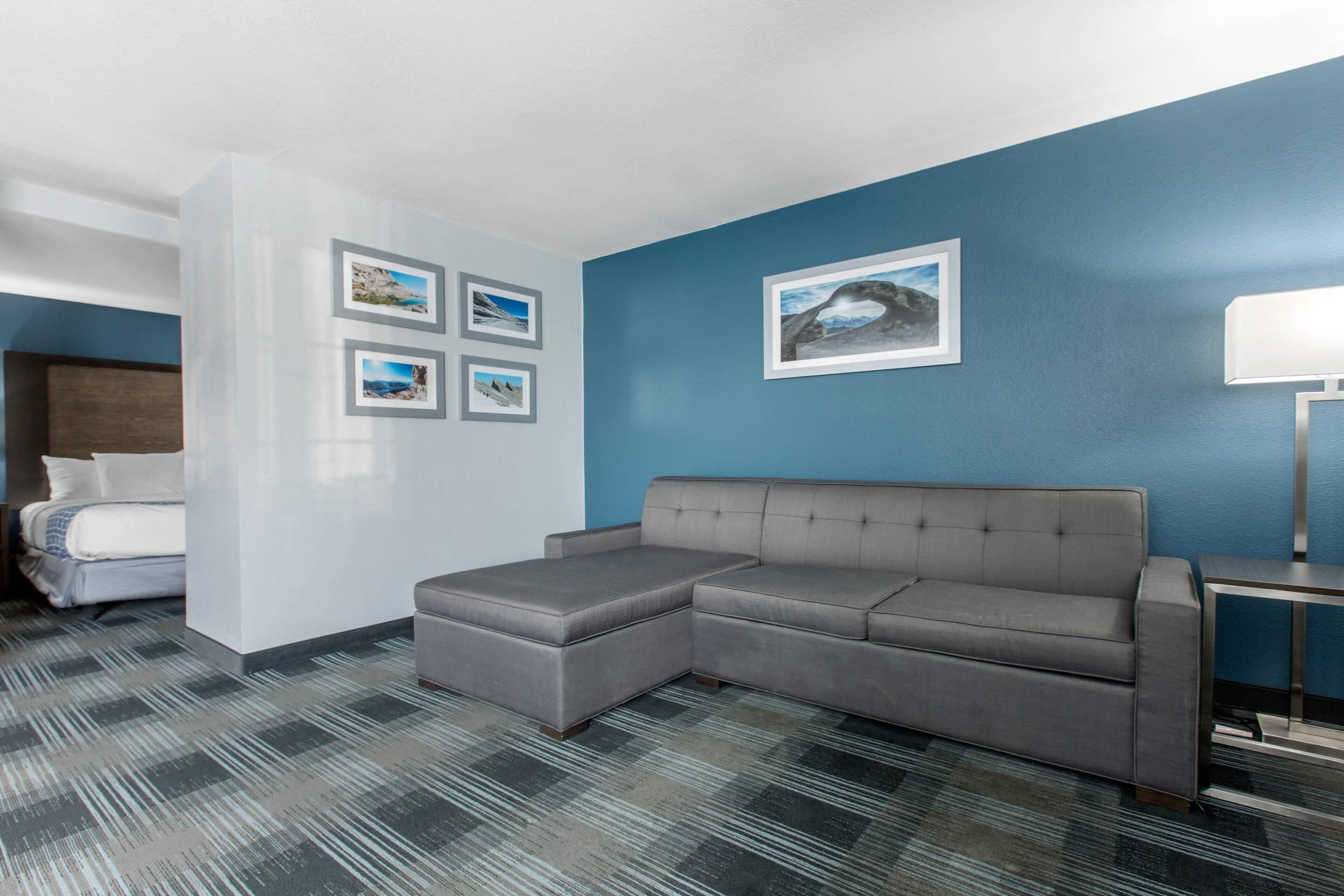 Ett modernt hotellrum med en grå, modulbaserad soffa, en säng med vita sängkläder, en blå accentvägg och inramade tavlor.