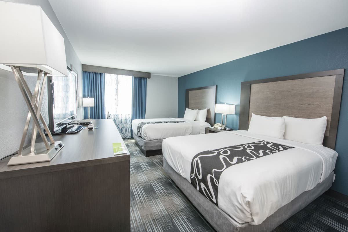 Une chambre d’hôtel avec deux lits, un bureau avec une lampe, un téléphone et une fenêtre avec des rideaux.