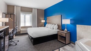 Hotelkamer met een bed, nachtkastjes, een bureau met stoel, bureaulamp, een kapstok en een raam met gordijnen, plus een badkamer met bad.