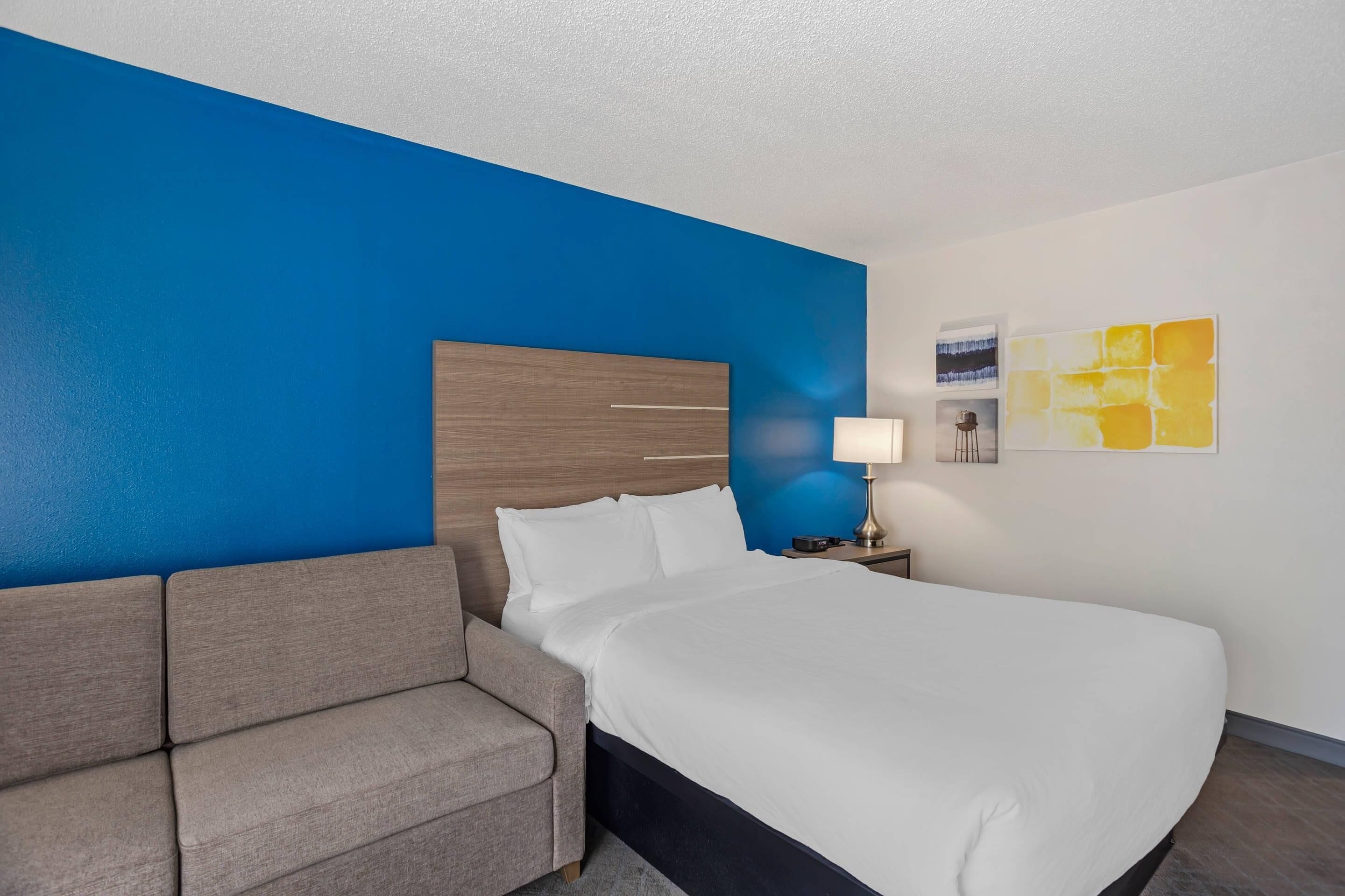 A hotel room with a bed, a sofa, a nightstand with a lamp, and a wall with a painting.