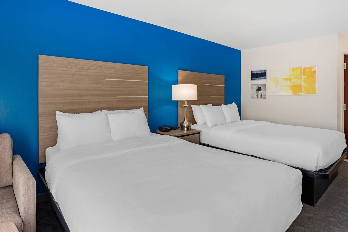 A hotel room with two beds, a nightstand with a lamp, and a chair.
