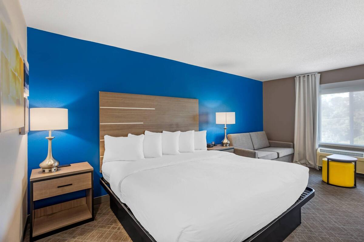 A hotel room with a large bed, two bedside tables with lamps, a sofa, a small yellow side table, and a window with curtains.