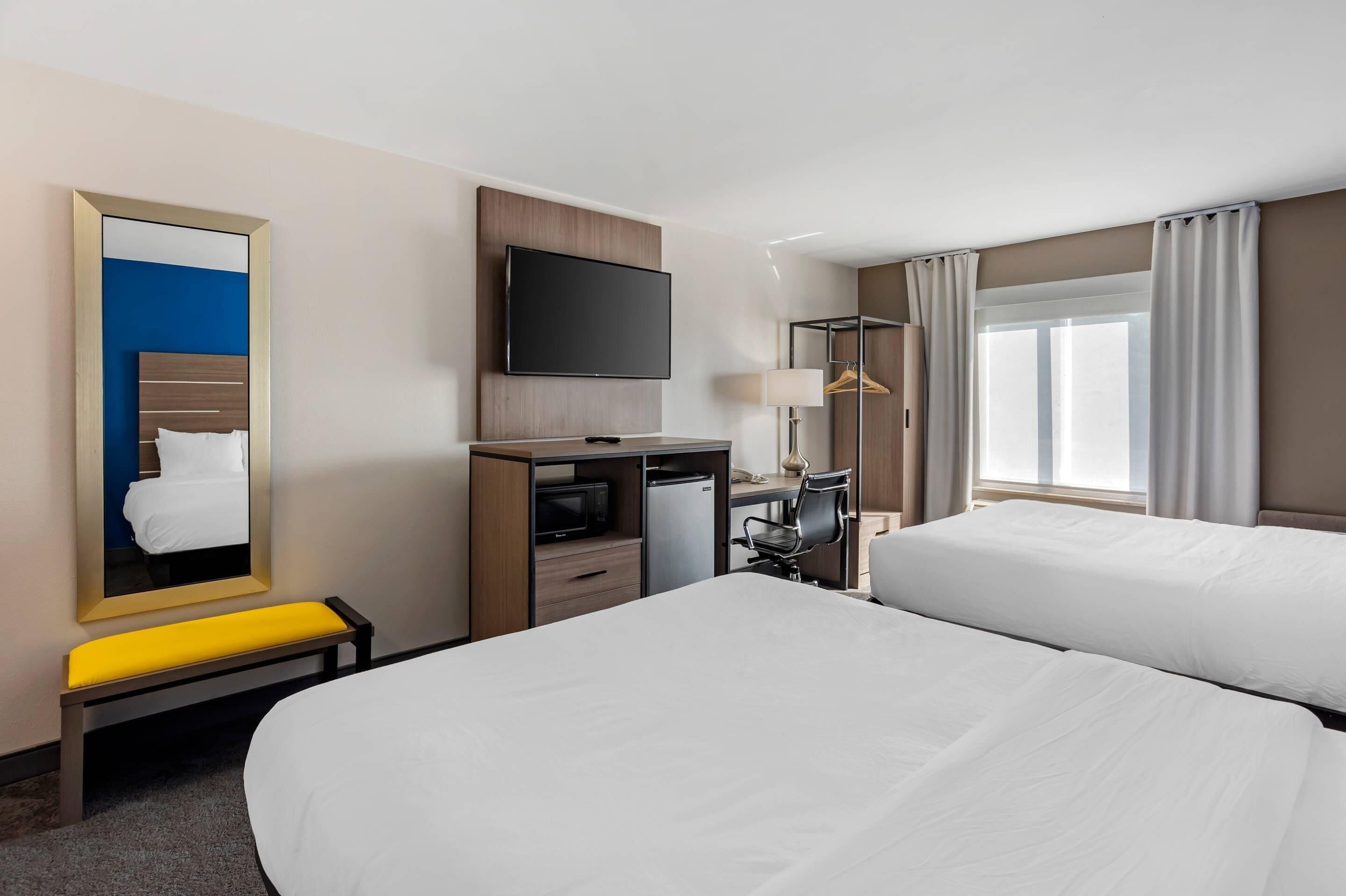 A hotel room with two beds, a TV, a desk with a computer, a lamp, and a window with curtains.