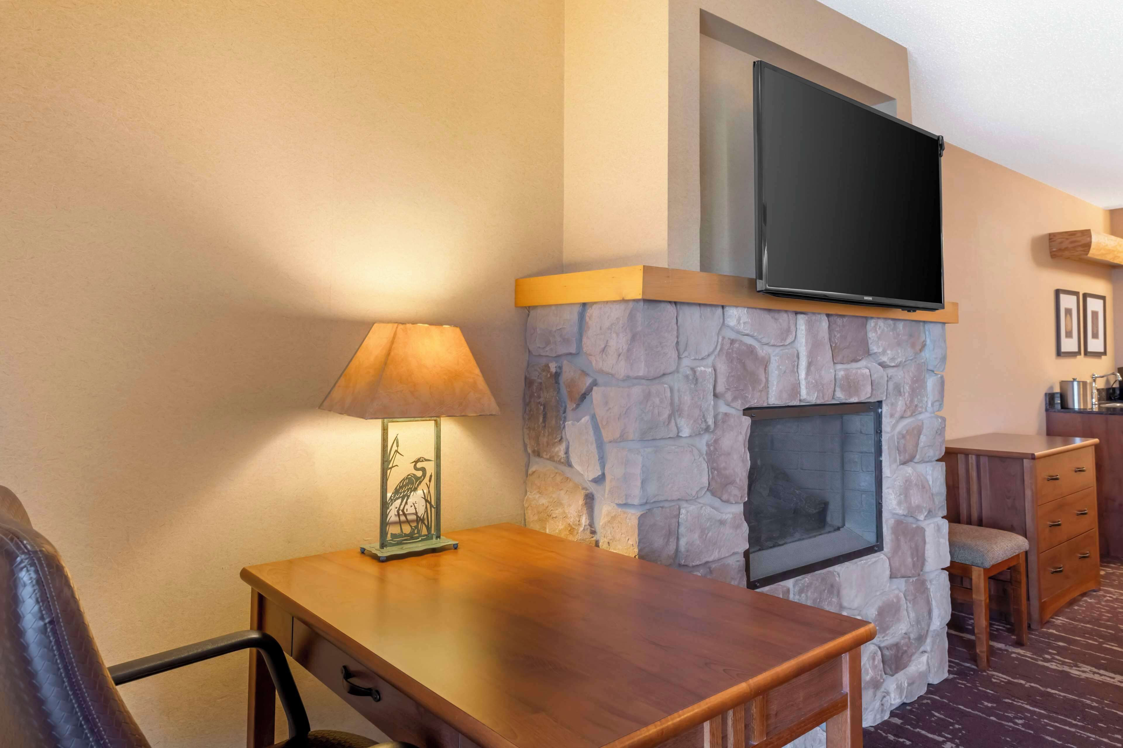 Una habitación con chimenea de piedra, un televisor de pantalla plana montado encima, una mesa de madera con una lámpara, y una zona de estar con una silla y una mesita.