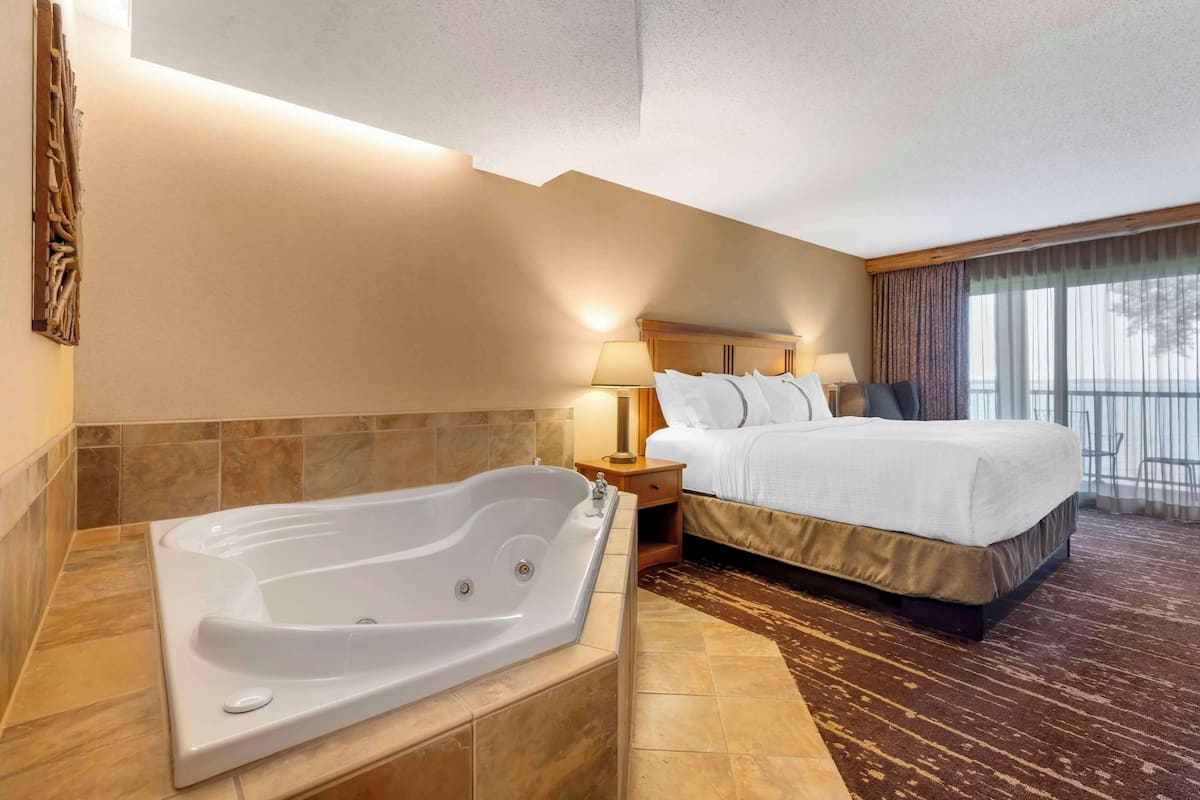 A hotel room with a large bathtub, a bed with white linens, a nightstand with a lamp, and a view of the outdoors through the window.