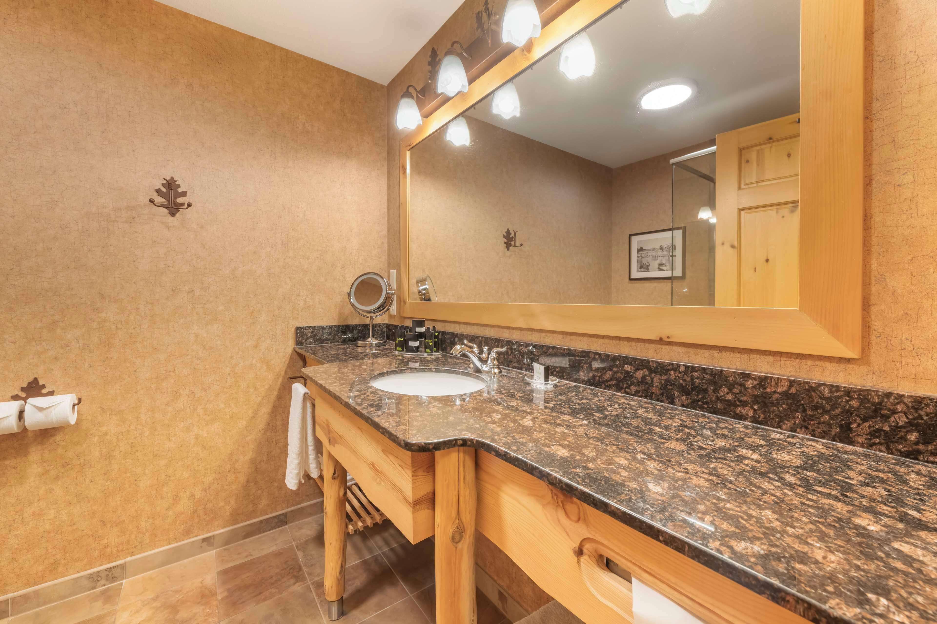 Un baño con lavamanos de encimera de granito, mueble de tocador de madera y un espejo grande con iluminación.