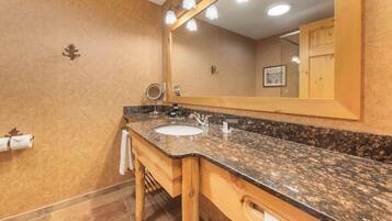 A bathroom with a granite countertop sink, wooden vanity, and a large mirror with lights.