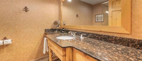 A bathroom with a granite countertop sink, wooden vanity, and a large mirror with lights.