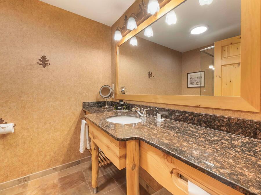 A bathroom with a granite countertop sink, wooden vanity, and a large mirror with lights.