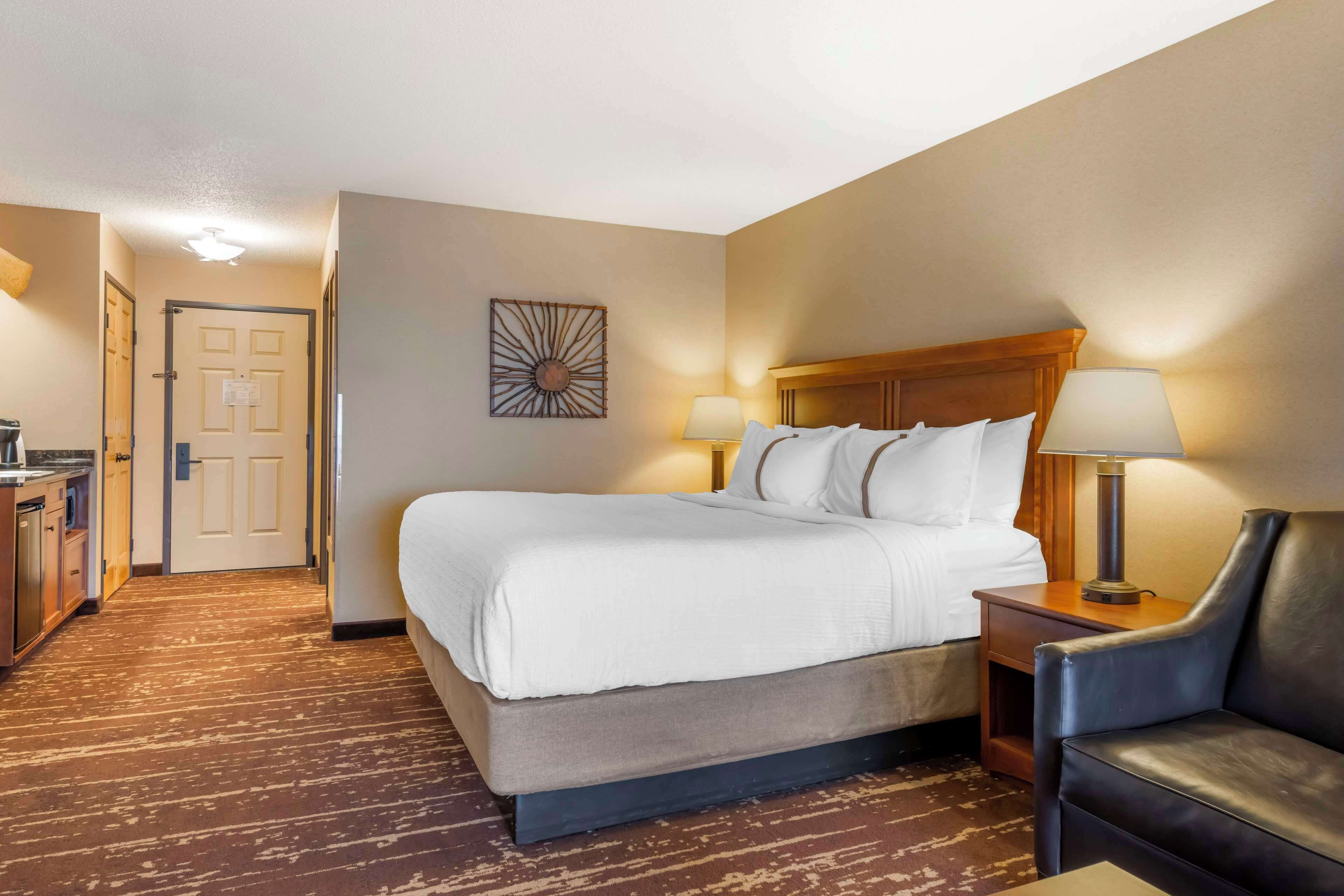 A hotel room with a large bed, a desk with a lamp, a chair, and a door leading to a hallway.