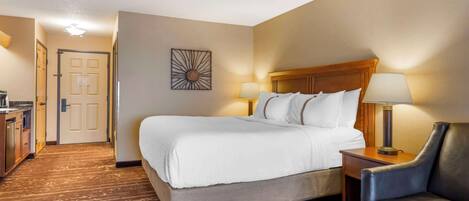A hotel room with a large bed, a desk with a lamp, a chair, and a door leading to a hallway.
