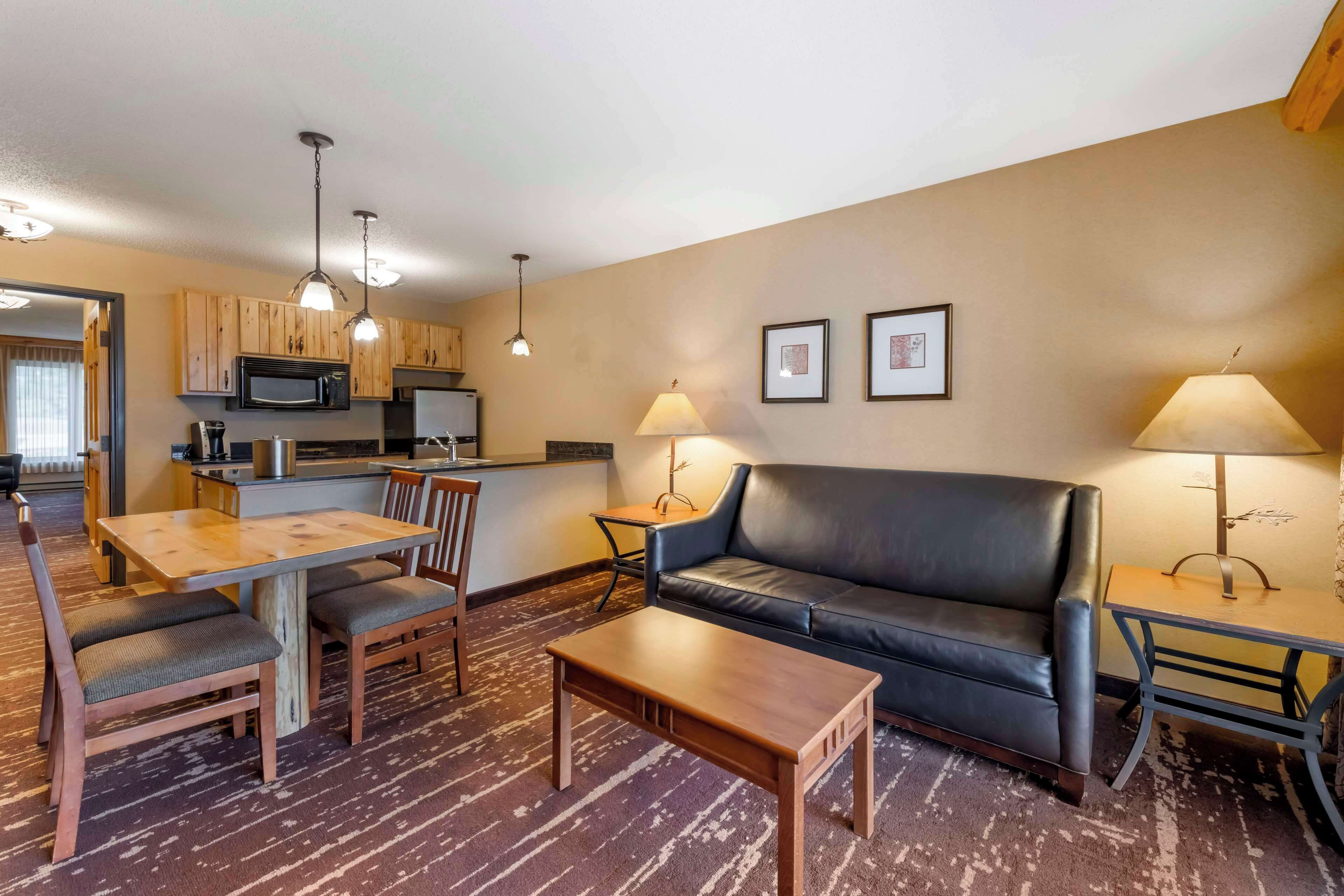 A modern living room with a wooden dining table, a black leather sofa, and a coffee table. There is a kitchen area with wooden cabinets and a microwave. Two lamps provide lighting, and the walls are decorated with framed pictures.